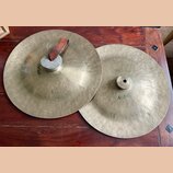 Dvě kovové činely v mosazné barvě s viditelným klepaním povrchu. Na levém číneli je nápis Meinl a připevněný hnědý kožený popruh. Oba nástroje vykazu…