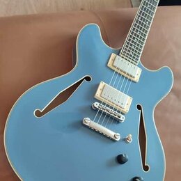 Modrá polodublová elektrická kytara s dvěma humbuckery a bílým lemováním kolem těla. Nástroj má dva černé ovládací knoflíky a stříbrný most s úpravou…