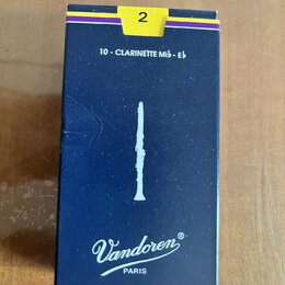 Modrá krabička s žlutým a fialovým pruhem, na které je bílá silueta klarnetu. Jsou zde nápisy „2“, „10 - CLARINETTE Mib - Eb“ a logo „Vandoren PARIS“…