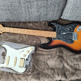Elektrická kytara s tělem v provedení sunburst a hnědým hřebenatým pickguardem. Na hlavě je viditelné logo Washburn. Vedle kytary leží v plastovém ob…