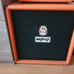Kytarový reproduktor v oranžovém potahu s černou textilní mřížkou. Na mřížce je umístěno bílé obdélníkové logo s nápisem ORANGE a nad ním barevná nál…