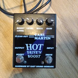 Černý kytarový efekt s nápisy CARL MARTIN a HOT DRIVE'N BOOST. Na horní straně jsou čtyři ovládací knoflíky s popisky BOOST LEVEL, LEVEL, WAVE a GAIN…