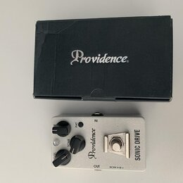 Kytarový efekt pedál stříbrné barvy s nápisy Providence a SONIC DRIVE. Pedál má tři černé ovládací knoflíky s popisky FAT, TONE a LEVEL. Vedle je umí…