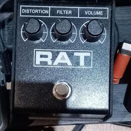 Černý efekt pedál s nápisem RAT v bílém obdélníkovém rámečku. Horní část obsahuje tři ovládací knoflíky s popisky DISTORTION, FILTER a VOLUME. Povrch…