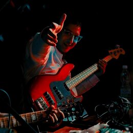 Fotografie zachycuje muže s červenou elektrickou basovou kytarou. Nástroj má červené tělo, bílý pickguard a světle hnědý hmatník. Na snímku nejsou vi…