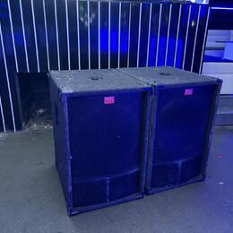 Dva černé, hranaté reproduktorové boxy s povrchem z tmavé textury. Na přední straně každého boxu je mřížka a červený obdélníkový štítek s bílým nápis…