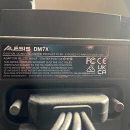 Fotografie detailně zobrazuje štítek s technickými údaji na černém plastovém zařízení. Na štítku je uveden název ALESIS DM7X a varování CAUTION: DO N…