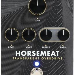 Černý efekt pedál s bílým vlnovitým vzorem na povrchu. Na horní části jsou čtyři černé otočné ovladače s popisky GAIN, LEVEL, VOICE, TREBLE a BASS. V…