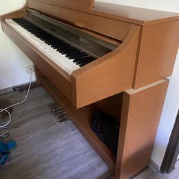Digitální piano v dřevěném korpusu s hnědým povrchem. Nástroj má bílé a černé klávesy a tři pedály v dolní části. Na povrchu nejsou vidět žádné značk…