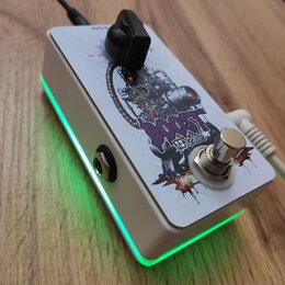 Bílý efekt pedál s černým ovládacím knoflíkem a stříbrným spínacím tlačítkem. Na horní straně je grafika s nápisem BOOST a menším textem pedals. Bočn…