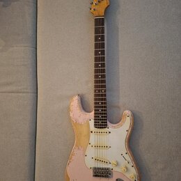 Růžová elektrická kytara typu Stratocaster s bílým pickguardem a hnědým hmatníkem. Lak na těle vykazuje výrazné opotřebení a odrolení v okrajích, kde…