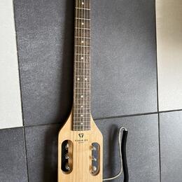 Světle hnědá elektrická kytara s krátkým tělem a dlouhým hnedým hmatníkem. Na těle je černý nápis TRAVELER GUITAR a nad ním logo ve tvaru trojúhelník…