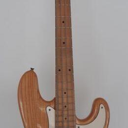 Elektrická basová kytara s přírodním dřevěným povrchem těla a hmatníkem. Na hlavě je viditelné logo "Fender". Instrument má bílý pickguard a čtyři st…