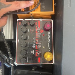 Bílý efekt pedál s červenými pruhy a nápisy BIG MUFF, deluxe a electro-harmonix. Zařízení má pět černých otočných ovladačů označených LEVEL, MUDS, SU…