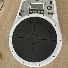 Bílý elektronický perkusní pad Roland HandSonic 10 s velkým černým gumovým playing polem. V horní části je ovládací panel s displejem, několika tlačí…