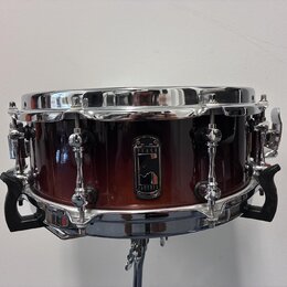 Kytarový snare drum s korpusem v červenohnědém přechodovém odstínu a chromovanými obručemi. Uprostřed korpusu je černý štít s bílým logem a nápisy BL…