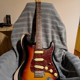 Elektrická kytara s trupem v barevnosti sunburst a hnědým hmatníkem. Kytara má červeně mramorovaný pickguard a na hlavě je viditelné logo Fender. Pov…