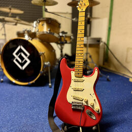 Červená elektrická kytara typu Stratocaster s bílým pickguardem a světle hnědým hmatníkem. Na hlavě kytary je viditelné žluté logo. Nástroj stojí na…
