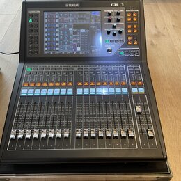 Černý digitální mixovací pult značky YAMAHA, model QL1. Horní část obsahuje barevný displej s ovládacími prvky, několik posuvníků a tlačítek. Spodní…