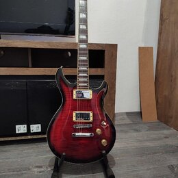 Elektrická kytara s červeným gradientem a černým okrajem, umístěná na černém stojanu. Na hlavě hmatníku je bílý nápis Cort. Nástroj má dva humbuckery…