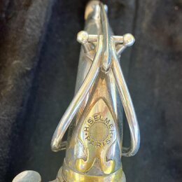 Detailní pohled na horní část saxofonu v stříbrném povrchu s mosaznými prvky. Na těle nástroje je vidět kulaté logo s nápisem H. SELMER PARIS. Povrch…