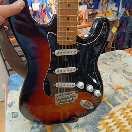 Elektrická kytara s trupem v barevnosti sunburst a černým pickguardem. Na kytaru je nasazený hnědý popruh a jsou vidět tři snímače typu single-coil.…