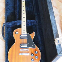 Elektrická kytara s trupem ve tvaru Les Paul v žlutohnatém plamenovém provedení, umístěná v černém tvrdém kufru. Na hlavě hmatníku je nápis Ovation a…