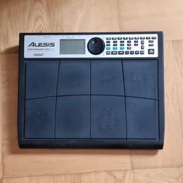 Černý elektronický bicí pad značky ALESIS, model PERFORMANCE PAD PRO. Zařízení obsahuje mřížku z gumových políček a ovládací panel s displejem, tlačí…