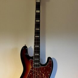 Čtyřstrunná basová kytara s korpusem v barevném přechodu od žluté přes oranžovou k černému. Nástroj má hnědý pickguard s červenými vločkami a na hlav…