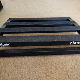 Černý kovový pedalboard s mřížkovou konstrukcí a vložkami z černého textilního popruhu. Na přední hraně je bílý nápis pedaltrain® a pod ním menší tex…