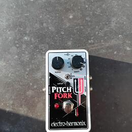 Kytarový efekt pedál v bílém kovovém krytu s černým ovládacím panelem. Na pedálu jsou nápisy PITCH FORK a electro-harmonix, dva černé otočné knoflíky…