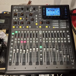 Digitální mixážní pult černé barvy s nápisem X32 PRODUCER a logem Behringer. Zařízení obsahuje displej, zahlreiche posuvníky, tlačítka a otočné ovlad…