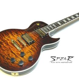Elektrická kytara s tvarem Les Paul, s plameníkovým topem v hnědé barvě a zlatým hardwarem. Na těle jsou dva humbuckery, čtyři ovládací knofíky a pře…