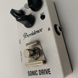 Bílý efekt pedál s nápisy Providence a SONIC DRIVE. Na horní straně jsou dva černé otočné knoflíky, jedno malé tlačítko s nápisem FAT a jeden malý šr…