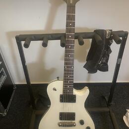 Bílá elektrická kytara s tmavým hmatníkem umístěná na černém stojanu. Na hlavě kytary je nápis "PRS Tremonti". Tělo má dva humbuckery, tři ovládací k…