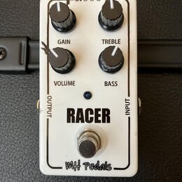 Bílý efekt pedál s nápisy RACER a MH pedals. Obsahuje čtyři černé ovládací knoflíky s popisky GAIN, TREBLE, VOLUME a BASS, vstup INPUT, výstup OUTPUT…