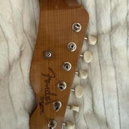 Detail dřevěné hlavy elektrické kytary v přirozené barvě s bílými ladicími kolíky. Na dřevě je vidět nápis Fender a pod ním text TELECASTER STRATOCAS…