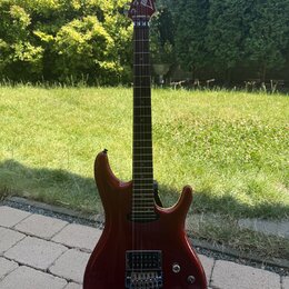 Elektrická kytara s červeným lesklým povrchem korpusu a hnědým hnedým hmatníkem. Na hlavě je bílý nápis Ibanez. Nástroj má dva humbuckery, kobylku s…