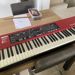 Červený digitální klavír s bílými a černými klávesami. Na horním panelu je nápis nord electro 5 v červeném rámečku a vlevo bílý text NEW FEATURES. Ov…