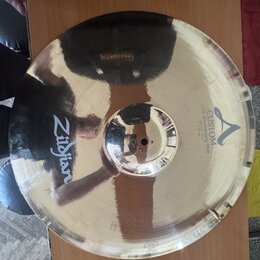 Zlatavý kovový činel s vyznačeným okrajem. Na povrchu je černý nápis Zildjian a logo A s textem CUSTOM 20th Anniversary Ride MEDIUM Thin 22". Povrch…