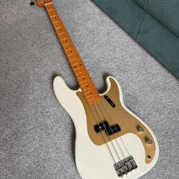 Bílá elektrická basová kytara s hnědým hmatníkem a zlatovými plastovými kryty. Na hlavě je nápis Fender. Nástroj má dva snímače, tři ovládací knoflík…