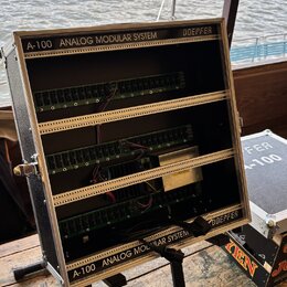 Stříbrný transportní kufr s černým vnitřkem, který obsahuje modulární syntetizátor. Na horní a spodní liště je bílý nápis A-100 ANALOG MODULAR SYSTEM…