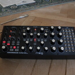 Černý analogový polyrytmický syntetizátor s nápisy SUBHARMONICON a moog. Přístroj obsahuje řadu bílých otočných ovladačů, červených podsvícených tlač…