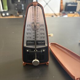 Hnědý mechanický metronom s bílým úzkým úželem a černým spodním panelem. Na přístroji jsou nápisy "Wittner METRONOM", "taktell PICCOLO" a označení te…