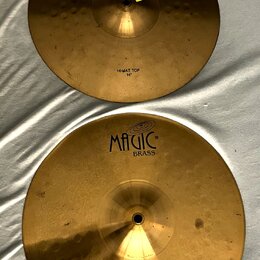 Dvě mosazné činely v zlatavé barvě. Oba mají nápis MAGIC BRASS a jsou označeny jako HI-HAT TOP 14" (horní) a HI-HAT BOTTOM 14" (dolní). Povrch činelů…