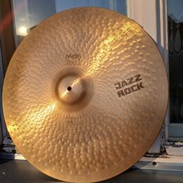 Kruhová mosazná činely s kovaným povrchem a viditelnými rýhami. Na povrchu jsou nápisy „paiste 2002“, „JAZZ ROCK“ a v dolní části „HEAVY RIDE“. Povrc…