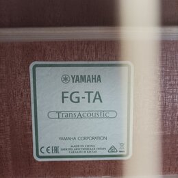 Detail štítku uvnitř akustické kytary. Na bílém pozadí s zeleným rámem je uveden nápis YAMAHA, model FG-TA a označení TransAcoustic v oválu. Dole je…