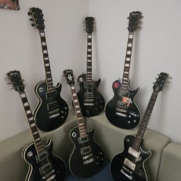 Šest černých elektrických kytar typu Les Paul s bílými bindingy. Na hlavách některých nástrojů je viditelný nápis Epiphone. Kytary mají různé konfigu…