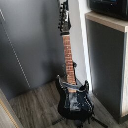 Černá elektrická kytara s hnědým hmatníkem umístěná na stojanu. Na hlavě je bílý nápis Ibanez. Kytara má tři snímače, černý pickguard a připevněný še…