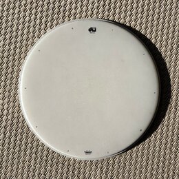 Bílá kruhová bubenová koží v kovovém obruči. V horní části je černé logo „dw drums“ a nápis „coat batter“. Dole se nachází modré logo „Remo“ a text „…
